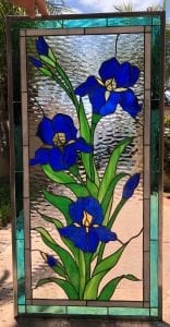 A rectangular stained glass panel depicting three vibrant blue iris flowers with green leaves and unopened buds. The irises have subtle yellow accents in their centers. The background glass is clear and heavily textured, resembling rippling water. The panel is bordered by lead came and features outer sections of clear, white, and streaky teal-blue art glass.