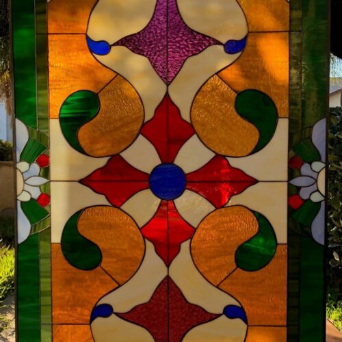 A rectangular, leaded stained glass privacy panel with a symmetrical, ornate Victorian-style design. It features a central blue roundel surrounded by radiating red, amber, and textured purple glass, all set against a creamy white opaque background. The panel is bordered by solid green glass and includes delicate white and green floral motifs in the corners and along the top and bottom edges.
