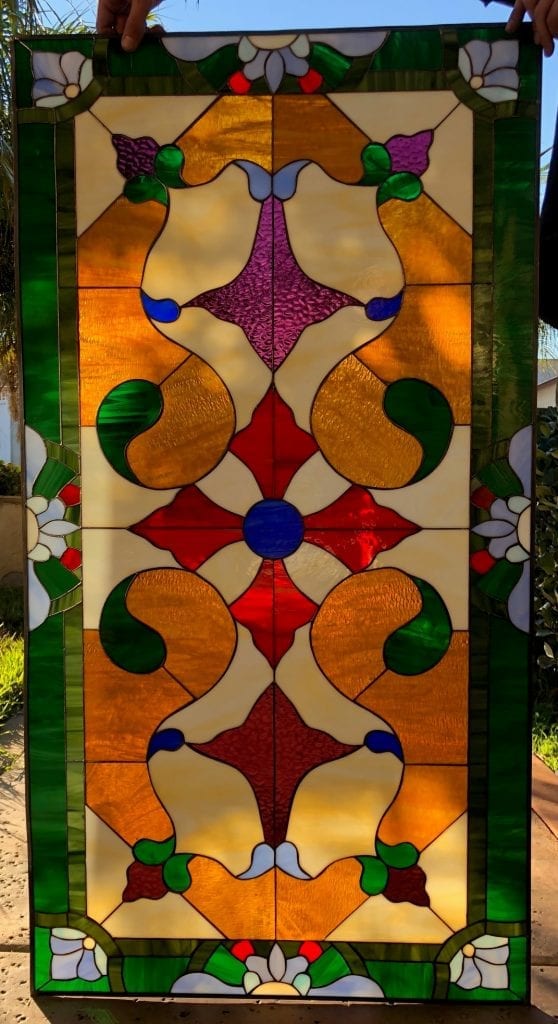 A rectangular, leaded stained glass privacy panel with a symmetrical, ornate Victorian-style design. It features a central blue roundel surrounded by radiating red, amber, and textured purple glass, all set against a creamy white opaque background. The panel is bordered by solid green glass and includes delicate white and green floral motifs in the corners and along the top and bottom edges.