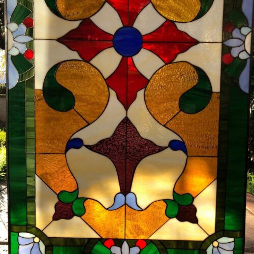 A rectangular stained glass panel featuring an ornate, symmetrical design. A central red cross with a blue circle is surrounded by four textured golden scroll-like motifs and cream-colored glass. The panel is framed by a rich dark green border with delicate light green and white floral accents.