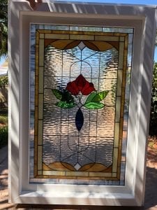 A rectangular stained glass insulated window unit featuring a central red rose with green leaves and a dark blue teardrop shape. The background is clear rippled textured glass, framed by an intricate border of amber, gold, and clear geometric panels, all encased in a modern white frame.