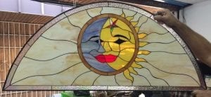 An arched leaded stained glass panel depicting a split sun and moon face. The left side shows a blue-grey crescent moon with a tranquil expression, while the right side shows a radiant yellow sun with a star detail and sun rays. Both celestial figures share vibrant red lips. The background features wavy, semi-translucent cream-colored glass, framed by a border of clear textured glass.