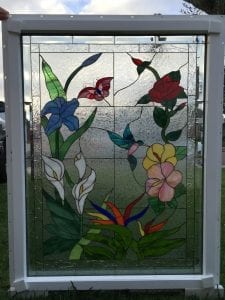 A rectangular stained glass window panel in a white frame, featuring a vibrant garden scene with white calla lilies, blue irises, a red rose, pink and yellow hibiscus-like flowers, and orange bird of paradise plants. A red butterfly and a teal hummingbird are shown among the flora, all set against a clear, textured privacy glass background.