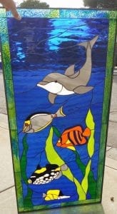 A vibrant rectangular stained glass panel featuring an underwater ocean scene. A gray dolphin with a white belly swims in the deep blue water at the top. Below, a diverse group of colorful tropical fish including a striped orange, a black and white spotted, a yellow and gray, and a small yellow fish are depicted. Lush green seaweed rises from the bottom, and the top portion shows lighter blue water with surface ripples. The panel is framed with a textured green border.