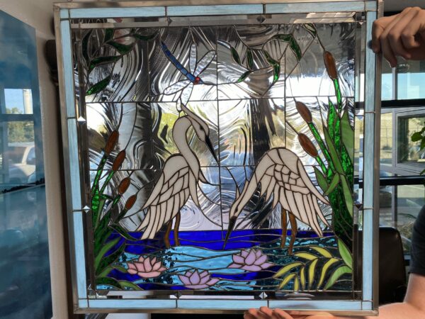 A rectangular leaded stained glass panel depicting a serene wetland scene. Two elegant white egrets stand in vibrant blue water, surrounded by pink and lavender water lilies, tall green cattails, and lush foliage. A small blue dragonfly with a red head hovers in the upper clear, ripple-textured glass background. The panel is framed with light blue glass accents and leaded borders.
