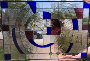 A rectangular stained glass panel with an abstract, geometric design. It features a central spiral motif with concentric arcs made of various clear textured and rippled glass. Royal blue curved and square glass pieces, deep ruby red sections, and small circular amber and green glass gems are incorporated throughout. The lead lines define the dynamic, flowing composition.