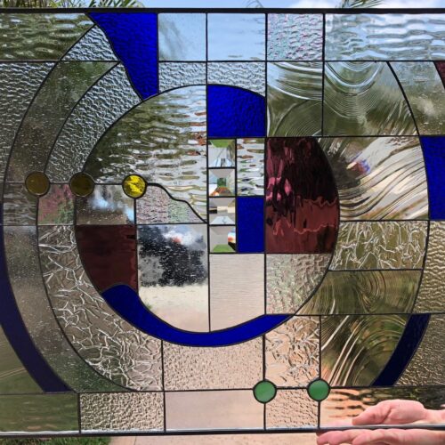 A rectangular stained glass panel with an abstract, geometric design. It features a central spiral motif with concentric arcs made of various clear textured and rippled glass. Royal blue curved and square glass pieces, deep ruby red sections, and small circular amber and green glass gems are incorporated throughout. The lead lines define the dynamic, flowing composition.