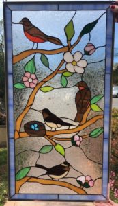 A rectangular stained glass panel featuring a vibrant spring scene with three birds perched on brown branches. One bird has a red breast, while two are brown with lighter bellies. Green leaves and delicate white and pink flowers adorn the branches. A bird's nest with three bright blue eggs is nestled on a branch. The background is clear, textured glass, framed by a blue border.