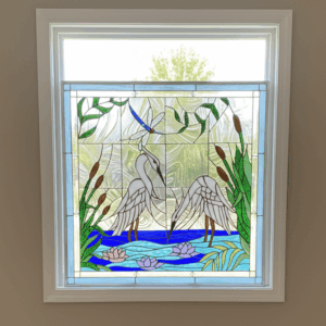 A rectangular, leaded stained glass window panel featuring two white herons wading in blue water with pink water lilies and green reeds. A blue dragonfly is depicted above one heron, set against a clear textured glass background.