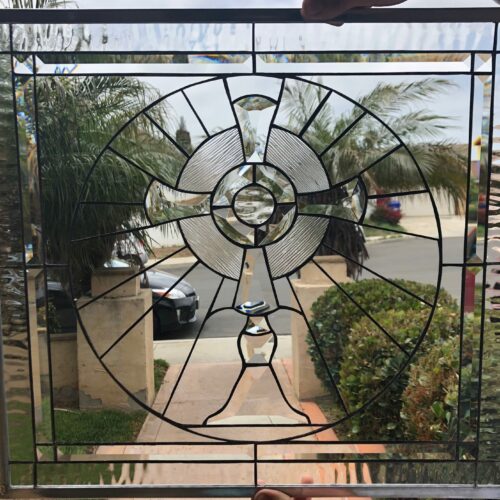 A rectangular, leaded stained glass panel with a central circular design composed of clear beveled, textured, and plain glass. The design features radiating lines, a central medallion, and a pedestal-like element at the bottom, all framed by a clear, textured border.