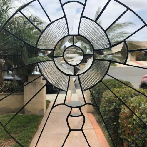 A circular leaded stained glass panel featuring a radiating geometric design composed of clear beveled glass and clear reeded textured glass, set against an outdoor view of palm trees and a sidewalk.