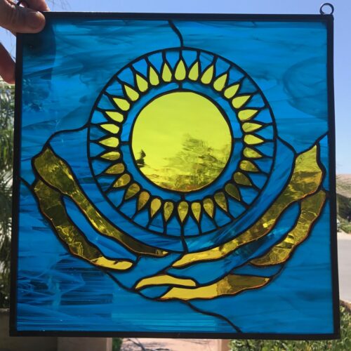 A square stained glass panel in vibrant blue and golden yellow, featuring the national emblem of Kazakhstan: a central golden sun with radiating teardrop-shaped rays, positioned above a stylized golden eagle or national ornament. The design is set against a textured azure blue glass background, bordered by dark leading and framed in black metal, reflecting bright sunlight.
