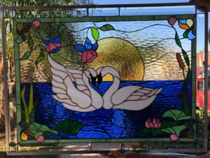 A rectangular leaded stained glass panel depicting two white swans with their necks gracefully curved to form a heart, floating on textured dark blue water. A large, textured golden sun/moon sets behind them, reflecting light. The panel is bordered by green glass and features vibrant pink and blue flowers, green foliage, lily pads, and cattails along the edges.