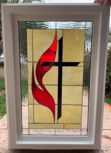 A rectangular stained glass panel in a white frame, depicting the United Methodist Church emblem with a vibrant red flame overlapping a black cross, set against a light amber background and bordered with clear textured glass.