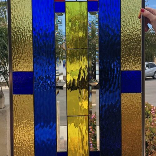 A rectangular stained glass panel with a vibrant abstract geometric design. It features a grid of various sized rectangles and squares in textured royal blue and golden yellow glass, interspersed with clear beveled and smooth clear glass, and punctuated by small red squares. The metal-framed panel is shown held outdoors against a blurred background of sky, palm trees, and a street.