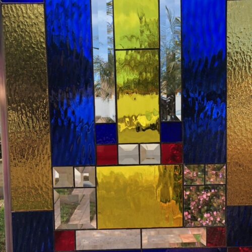 A rectangular stained glass panel featuring a bold, abstract geometric design with large vertical and horizontal blocks of textured cobalt blue, golden yellow, and smaller sections of smooth red and dark blue glass, interspersed with clear beveled rectangular and square pieces, set in a leaded frame.