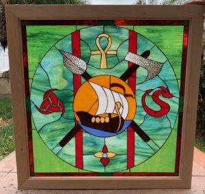 A square stained glass panel in a light wooden frame featuring a central circular design. Within the circle, a brown Viking longboat with white sails and golden accents floats on blue water against a golden sun. Behind the ship, a silver-bladed spear crosses a silver-bladed axe. Above the sun is a golden ankh, to the left a red Celtic knot, and to the right a red coiled serpent or dragon. The background inside the circle is a vibrant swirled green and blue glass, intersected by two bold red vertical stripes. The outer border area is a lighter, solid green.