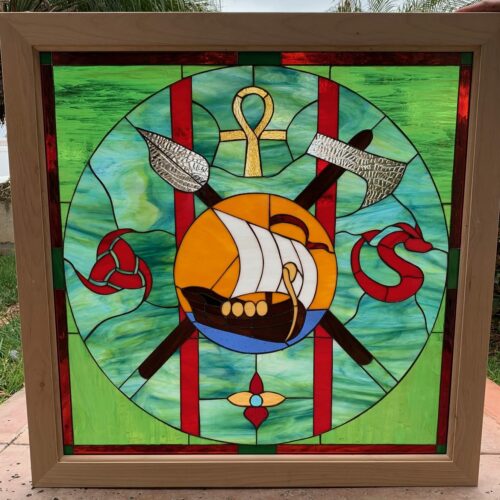 A square stained glass panel in a light wooden frame featuring a central circular design. Within the circle, a brown Viking longboat with white sails and golden accents floats on blue water against a golden sun. Behind the ship, a silver-bladed spear crosses a silver-bladed axe. Above the sun is a golden ankh, to the left a red Celtic knot, and to the right a red coiled serpent or dragon. The background inside the circle is a vibrant swirled green and blue glass, intersected by two bold red vertical stripes. The outer border area is a lighter, solid green.