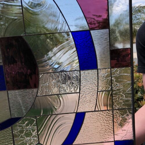 A rectangular leaded stained glass panel featuring a modern abstract design. The panel combines clear textured glass (including wavy, rippled, and crinkled patterns) with bold sweeping arcs of royal blue and deep plum glass. Two small green circular pieces are visible in the lower left. The transparent and translucent glass allows light and an outdoor background to show through, highlighting the varied textures and colors.