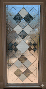A vertical rectangular stained glass panel featuring an interlocking diamond pattern crafted from clear, textured, white, and dark gray glass, framed in white.