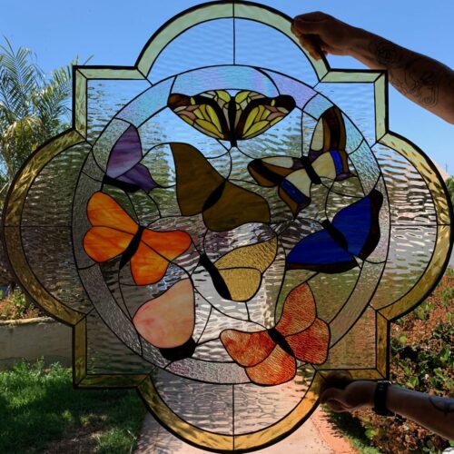 An ornate, quatrefoil-shaped stained glass panel featuring a vibrant central medallion with eight multicolored butterflies in shades of yellow, brown, purple, orange, blue, and peach. The background is clear, textured ripple glass, and the outer border is a golden-amber textured glass. The panel is held against a bright outdoor garden background, with hands visible at the top and bottom.
