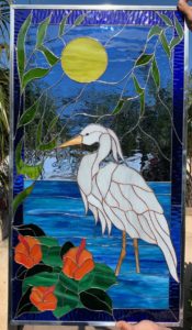 A rectangular stained glass panel featuring a majestic Great Egret standing gracefully in vibrant blue water. A bright yellow sun shines in the upper left, framed by delicate green leafy branches extending across the top. Three vivid orange-red hibiscus-like flowers with dark green leaves are prominent at the bottom left. The egret is meticulously rendered in textured white glass with a long yellow beak and slender brown legs. The water includes various shades of blue and clear textured glass, creating a serene ripple effect. The entire artwork is bordered by a deep blue glass frame and encased in a silver metal frame, suggesting it is a leaded stained glass piece.
