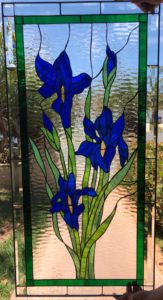 A rectangular stained glass panel depicting a bouquet of five vibrant blue irises with yellow centers, green stems, and leaves. The floral design is bordered by an inner dark green band and an outer clear beveled edge, set against a clear, rippled background glass.