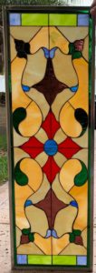 A rectangular leaded stained glass panel featuring a symmetrical, ornate design. It showcases a central red quatrefoil with a blue roundel, surrounded by flowing amber, brown, and green organic shapes, accented with small blue teardrops. The top and bottom borders include green and light blue rectangular pieces, all encased in a dark metal frame.