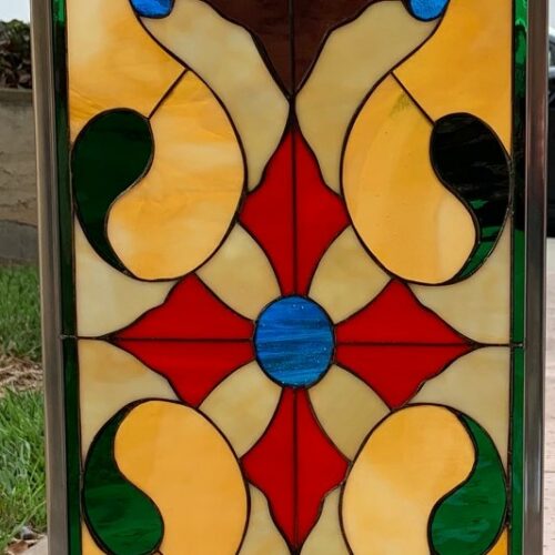 A rectangular leaded stained glass panel featuring a symmetrical, ornate design. It showcases a central red quatrefoil with a blue roundel, surrounded by flowing amber, brown, and green organic shapes, accented with small blue teardrops. The top and bottom borders include green and light blue rectangular pieces, all encased in a dark metal frame.