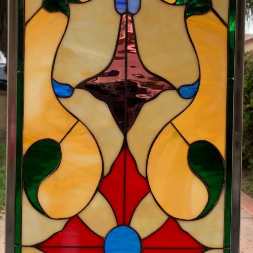 Rectangular stained glass panel with a symmetrical Art Nouveau-inspired design featuring amber, red, blue, green, and purple glass, centered around a red cross and blue medallion, framed in metal.