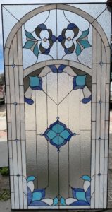 An arched leaded stained glass panel featuring abstract floral motifs in light and dark blues, gray, and white, set against a background of clear textured glass. Small blue glass jewels are embedded in the design.