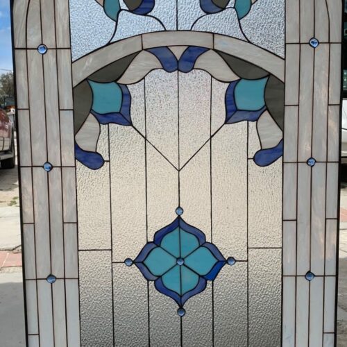 An arched leaded stained glass panel featuring abstract floral motifs in light and dark blues, gray, and white, set against a background of clear textured glass. Small blue glass jewels are embedded in the design.