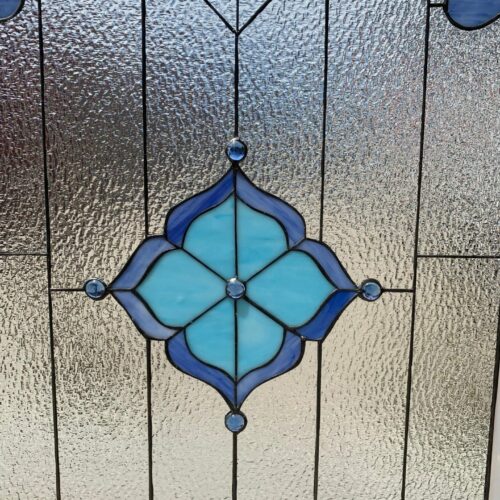 A close-up of a leaded stained glass panel featuring a symmetrical, stylized floral medallion in light blue and deep blue art glass, accented with clear blue glass jewels. The background consists of highly textured clear 'water glass' with dark lead lines, designed for privacy and light diffusion.
