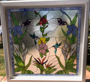 A rectangular stained glass window panel set in a white frame, depicting a vibrant and detailed garden scene. The central design features a red rose, blue irises, white calla lilies, and a Bird of Paradise plant. Two colorful hummingbirds with blue and green bodies are shown mid-flight near pink and yellow hibiscus-like flowers. Two butterflies with dark wings and blue accents are also present. The background glass is clear and textured, resembling hammered or ripple glass, allowing diffused light. A narrow blue stained glass border frames the main image, inside the white outer frame.