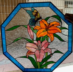 An octagonal stained glass panel depicting vibrant orange and pink lilies with green foliage, and a blue and yellow butterfly in flight, all set against a clear, textured background glass and framed by a luminous blue border.