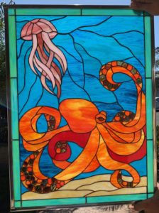 A rectangular leaded stained glass panel depicting a vibrant orange octopus with amber jewel suction cups, alongside a graceful pink jellyfish, against a swirling blue water background with a light sandy bottom.