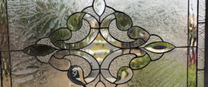 A close-up of a leaded stained glass panel showcasing a symmetrical, ornate central motif. The design is crafted from various clear textured glass types, including prominent glue chip glass and brilliant bevels, all connected by dark lead lines. The surrounding glass features a dense, organic frost or glue chip texture, providing privacy while allowing abundant light.