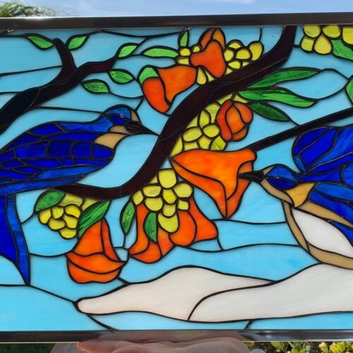 Rectangular stained glass panel depicting two vibrant bluebirds among branches with orange trumpet flowers and yellow clusters, set against a light blue sky. On the left, a blue bird is perched on a dark brown branch, facing right. On the right, another blue bird hovers or is in flight, its beak near an orange bell-shaped flower. The birds feature brilliant blue plumage with subtle tan and white accents. The branches are dark brown with green leaves, and the flowers are a rich orange, accompanied by clusters of yellow berries or small blooms. The entire panel is framed in a silver-toned metal border.