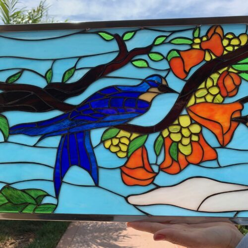 A rectangular stained glass panel in a silver-tone frame, depicting two bluebirds perched on a brown tree branch. The branch is adorned with green leaves, vibrant orange bell-shaped flowers, and clusters of yellow berries, all set against a light blue sky background.