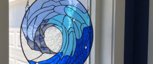 A circular leaded stained glass panel depicting a stylized breaking ocean wave, rendered in various shades of blue and clear textured glass, with sparkling clear glass gems.
