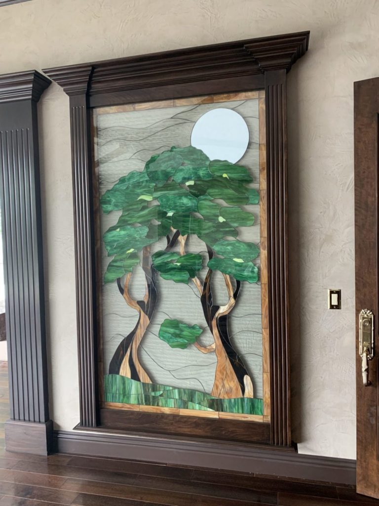 A large, rectangular stained glass art panel depicting a tranquil moonlit grove. Two stylized trees with vibrant green leaves and warm brown trunks stand against a wavy gray background. A bright white full moon is prominently featured behind the trees in the upper right. The panel is encased in an ornate, dark wood frame with fluted columns, installed within an interior wall.