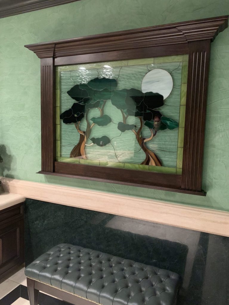 A rectangular stained glass panel depicting a moonlit scene with two dark green trees against a textured light green sky, featuring a large pale moon in the upper right. The panel is enclosed in a dark, fluted wooden frame.