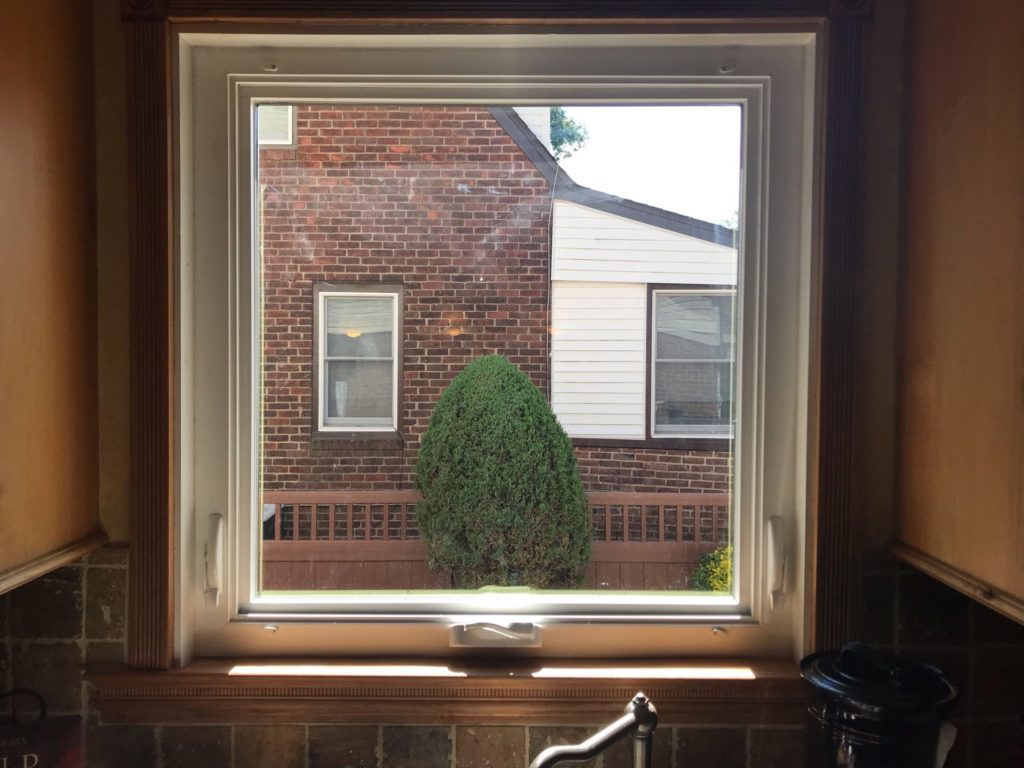 A rectangular clear and textured leaded stained glass panel, designed for privacy, installed within a kitchen window. It obscures the view of a neighboring brick house and shrub while allowing diffused natural light, featuring subtle beveled accents.