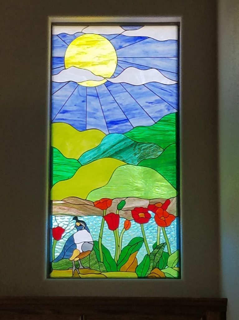 A rectangular stained glass panel depicting a vibrant nature landscape. In the upper portion, a large, bright yellow sun with radiating light lines shines through white clouds in a blue sky. Below the sky, rolling green hills of various shades are visible. The lower section features a blue, white, and brown quail-like bird standing on the left, next to several red poppies with green stems and leaves, all set against a light blue, watery background.