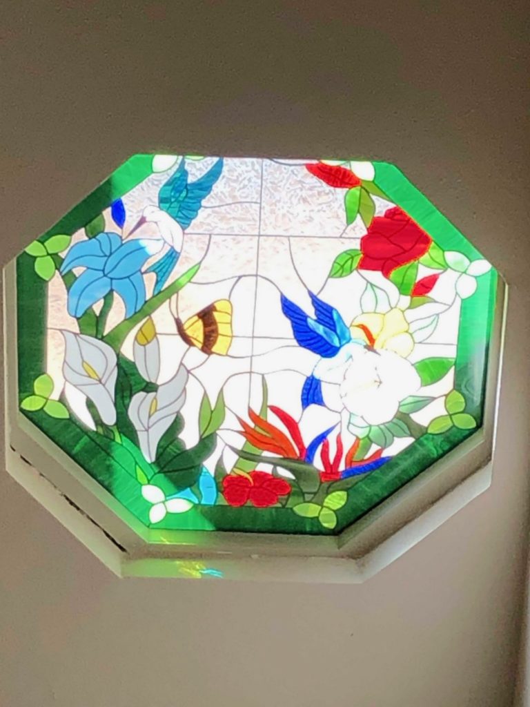An octagonal leaded stained glass panel depicting a vibrant tropical scene. It features a blue hummingbird, a yellow butterfly, white calla lilies, red roses, and orange bird of paradise flowers, all against a clear textured glass background with a green border.