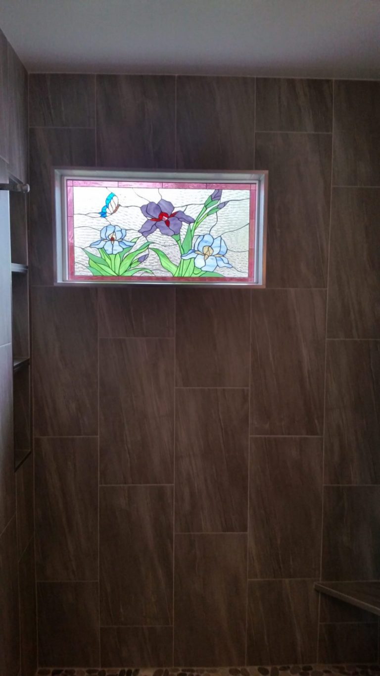 A rectangular stained glass panel installed high on a dark brown tiled shower wall. The panel features a vibrant design of three iris flowers (two light blue, one purple) with green leaves and stems, and a blue and white butterfly, all set against a clear, textured glass background. It has a thin light-colored frame with a red inner border.