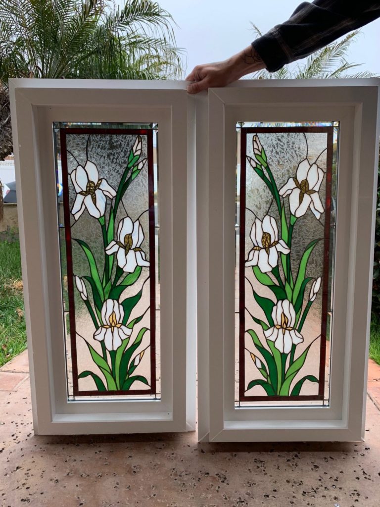 A pair of rectangular insulated window units, each containing a leaded stained glass panel depicting three white irises with golden-yellow centers, green stems, and leaves. The floral design is set against a clear, textured background and framed by a deep red border. The units are encased in thick white frames. A hand holds one panel from the top, against an outdoor background with palm trees.
