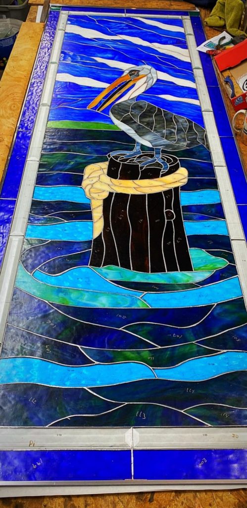 A rectangular leaded stained glass panel featuring a gray and white pelican with an orange beak, perched on a dark wooden piling wrapped with a yellow rope. The background depicts a blue sky with white wavy elements and a vibrant ocean with various shades of blue and green waves.