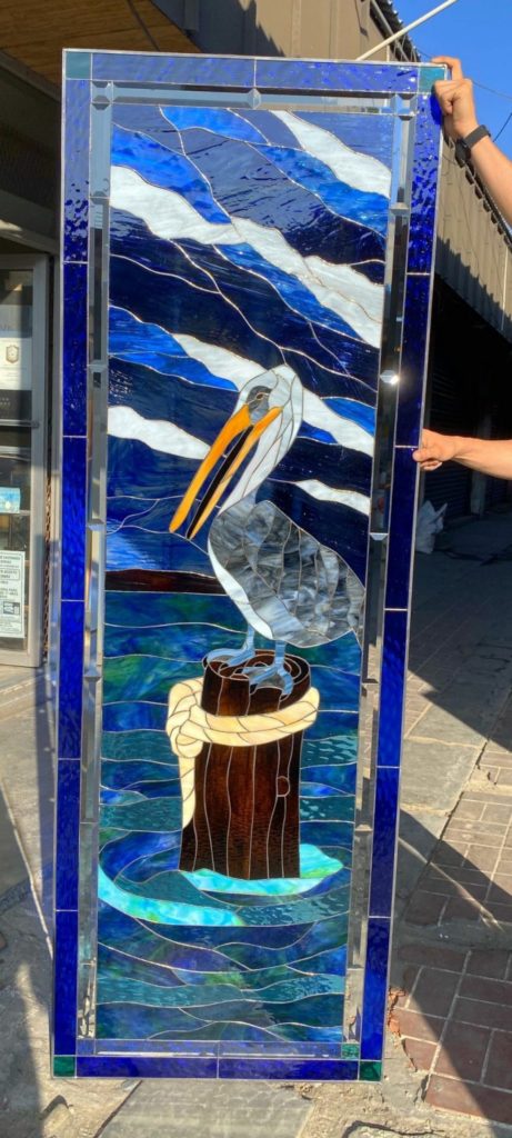 A rectangular, vertically oriented leaded stained glass panel depicting a gray pelican with an orange beak standing on a brown wooden piling with a white rope. The background features dynamic blue and white wavy glass, framed by a deep blue border with clear beveled accents.