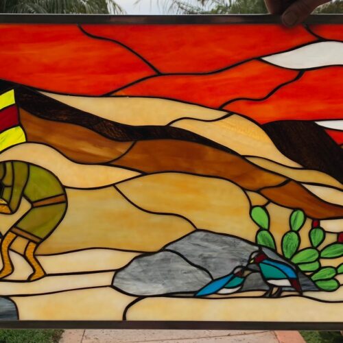 A rectangular stained glass panel depicting a vibrant desert sunset. The sky is fiery orange and red with white clouds over brown and tan hills. In the foreground, an Indigenous figure in olive green clothing and a feathered headdress plays a flute, watched by a green and blue striped lizard. To the right, a prickly pear cactus and a tall white agave flower rise from grey rocks, where two blue and red birds perch.